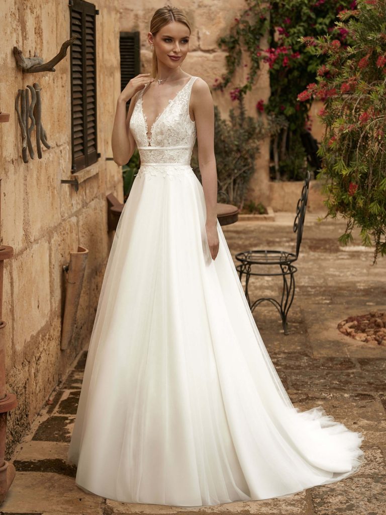 Photo shows a bride in a paved garden wearing Bianco Evento's Taylor A-line wedding dress. The bridal gown features a lace bodice with two narrow satin belts, one just under the bust, the other at the waist, and a plain tulle skirt.