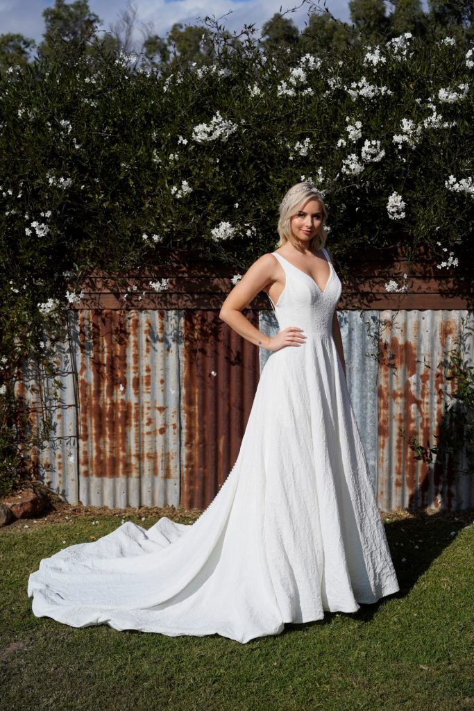 Photo shows a bride with her hand on her hip wearing Purple Fox's Cold November wedding dress, a plain ivory A-line bridal gown in a textured crepe with a v-neckline.