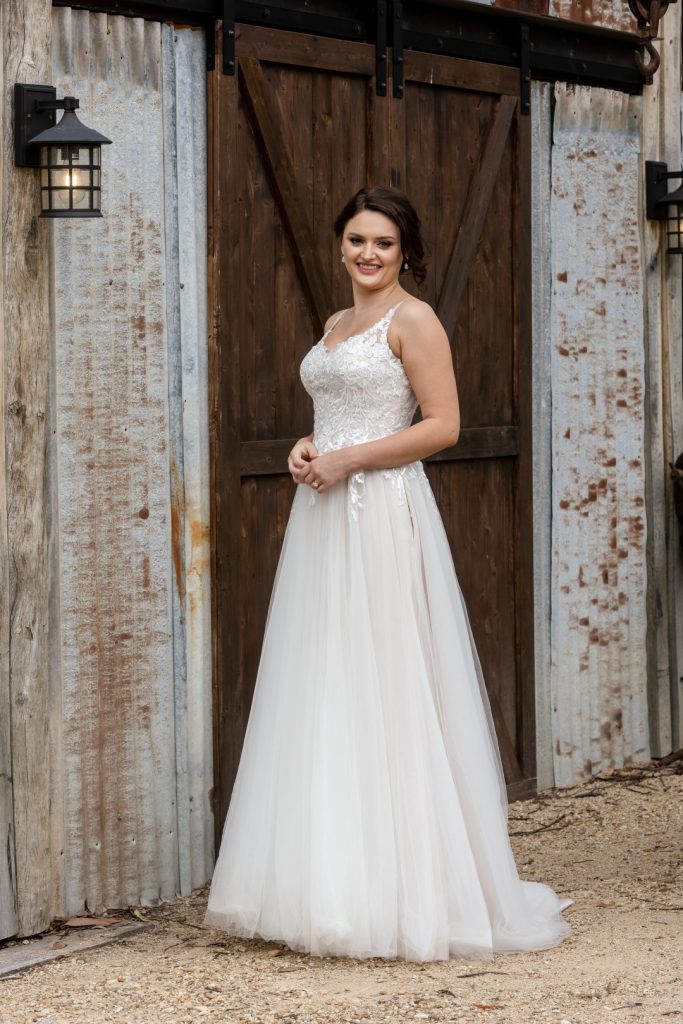 Photo shows a bride standing in front of a dark wooden door wearing Purple Fox's "Don't Stop" blush wedding dress, which features an ivory lace bodice with narrow straps and a plain tulle skirt. 