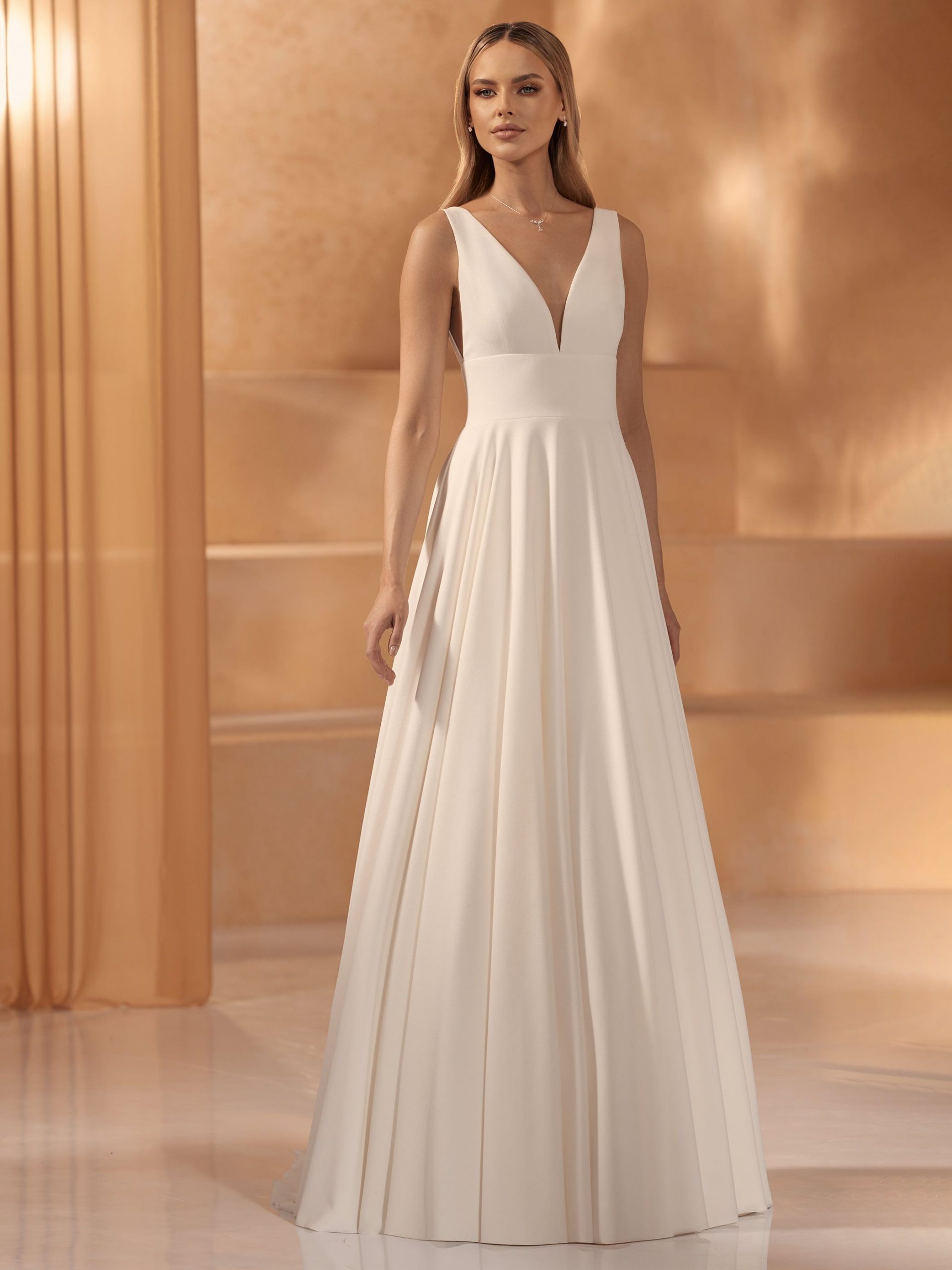 Photo shows a bride wearing a plain ivory crepe A-line wedding dress called Pola by Bianco Evento, featuring a low plunge neckline and wide waistband.