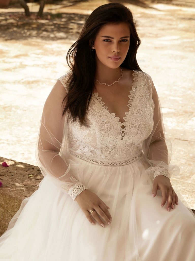 Photo shows a bride sitting on a wall wearing Bianco Evento's "Debora" A-line wedding dress. The ivory gown features a sequinned lace v-neck bodice, long tulle sleeves with lace cuffs, and a plain tulle skirt.