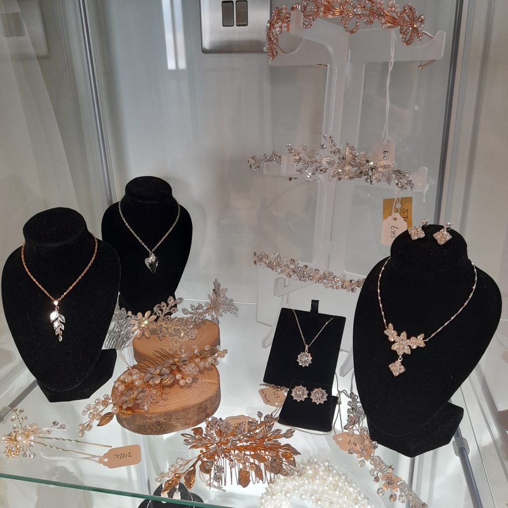 Photos shows a variety of necklaces, earrings, and hair accessories, in silver, gold, and rose gold, sitting in a glass cabinet.
