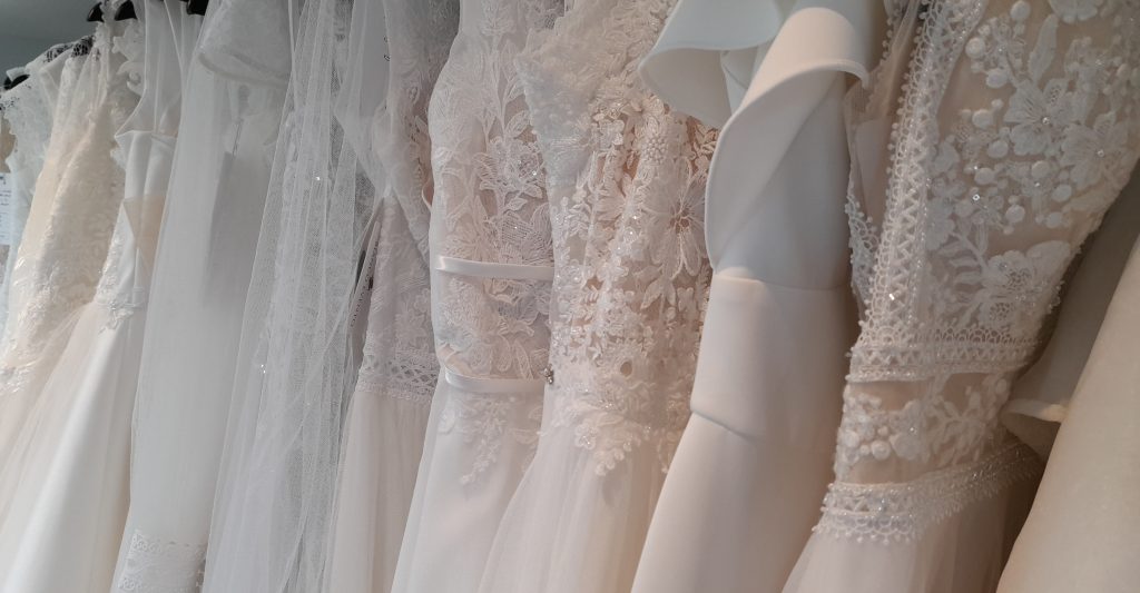Photo of several wedding dresses of lace, tulle, crepe, and chiffon, on a rail. Some of the bridal gowns are ivory, and some are blush. One has long sleeves.