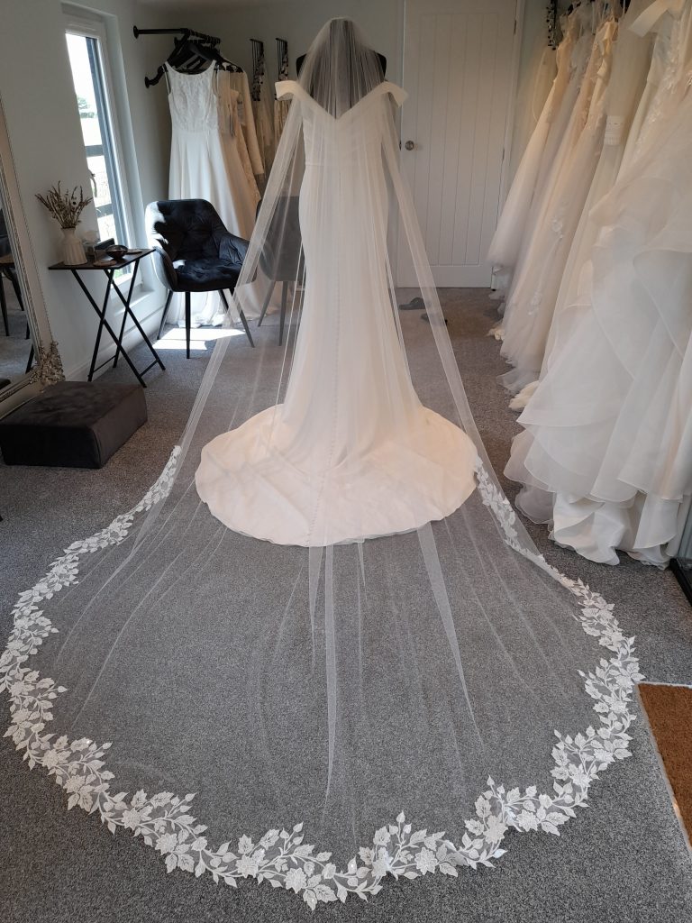Photo shows the inside of the bridal room with a mannequin dressed in a plain fitted bridal gown. There is a long veil with lace edging attached to the top of the mannequin.