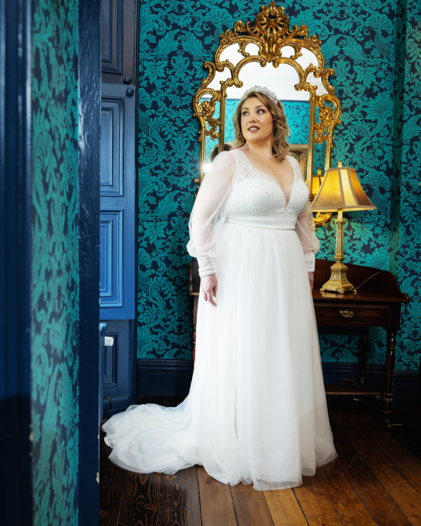 Photo shows a bride standing in a decadent room wearing an ivory wedding dress by Hilary Morgan. 40879 features a boho lace bodice with plunge neckline and long tulle sleeves with lace cuffs, a lace waistband, and plain tulle skirt.