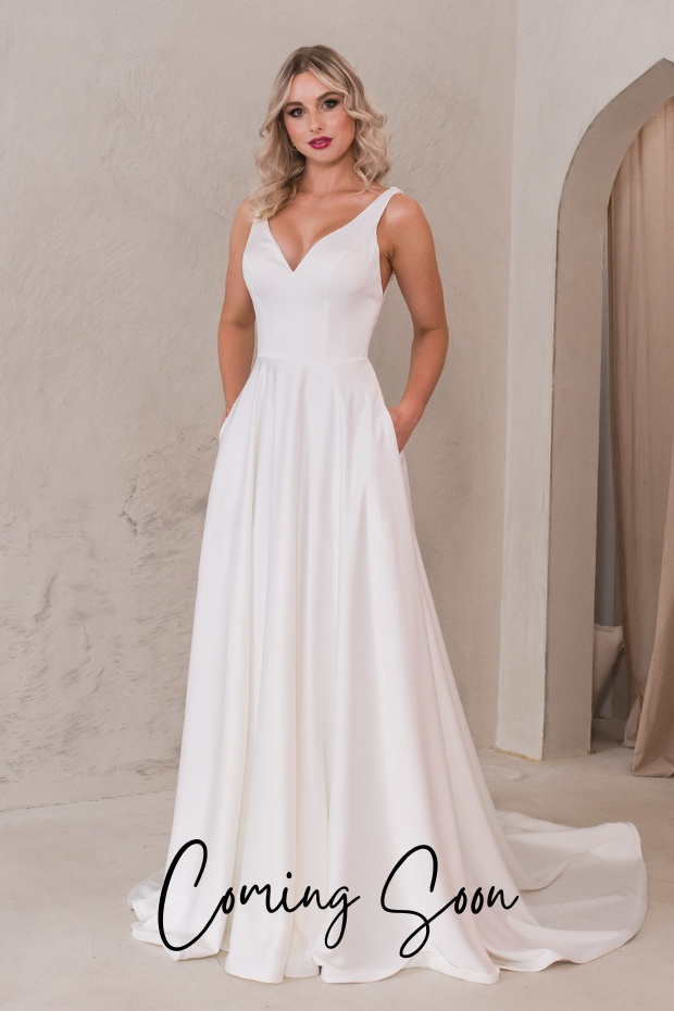 Photo shows a bride wearing an ivory satin A-line wedding dress by Purple Fox. December features a v-neckline, and plain skirt. The back bodice features 3 narrow straps with pearl beading and pearl buttons down the centre-back. This dress also has pockets!