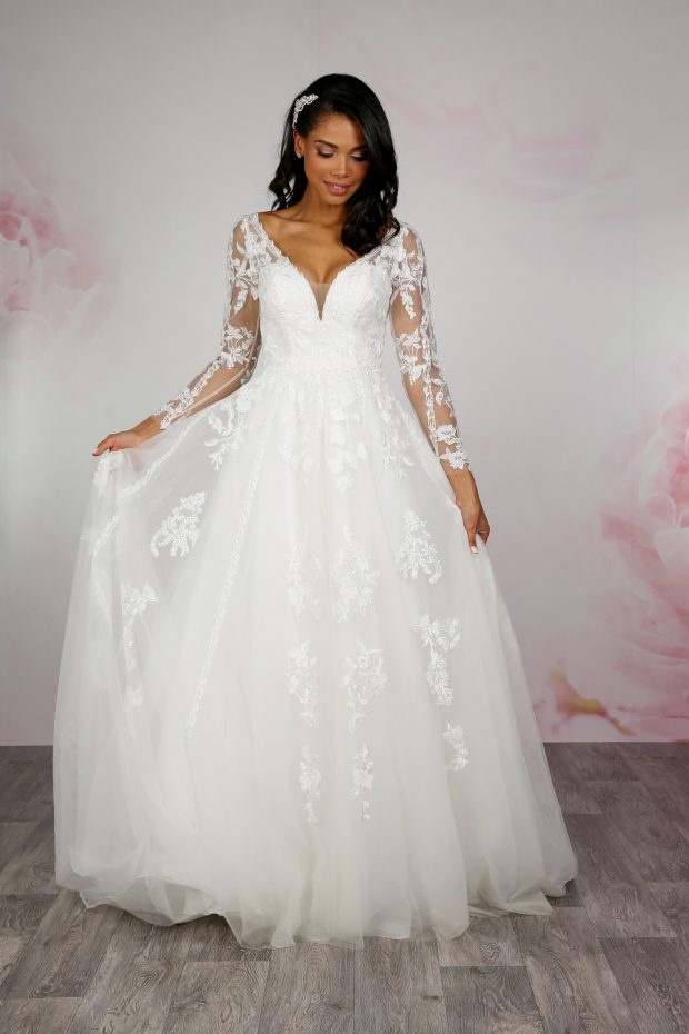 BL357 by Emma Bridals. An ivory A-line wedding dress with illusion bodice and long sleeves. The sleeves have been decorated with beautiful botanic lace appliques, echoed onto the bodice, and can be seen cascading down the skirt. Buttons have been added to the back of the dress, completing this bridal gown. Secured with a zip fastening and buttons at the back.