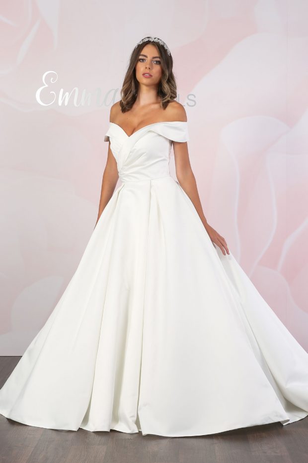 BL384 by Emma Bridals is an ivory satin off-the-shoulder ballgown with a flattering Bardot silhouette, pleated bodice and full skirt. Satin covered buttons feature down the centre-back seam all the way to the end of the train.