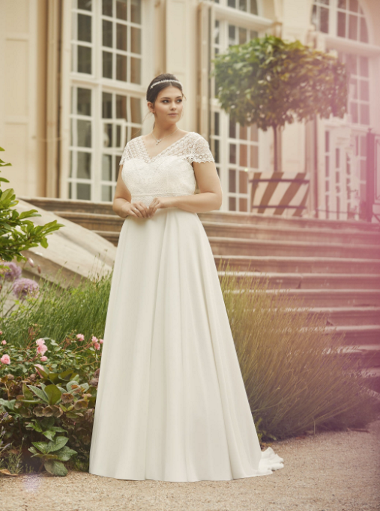 Photo shows a bride standing by a flower bed. She is wearing an ivory lace and chiffon wedding dress, called Margaret, by Bianco Evento. The bridal gown features a boho lace bodice with cap sleeves, over a plain sweetheart neckline, and a plain chiffon skirt.