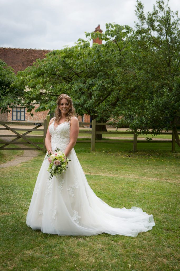 Photo of one of our real Lucky Sixpence brides. She is standing on grass, in front of a wooden fence and trees, holding her bouquet.