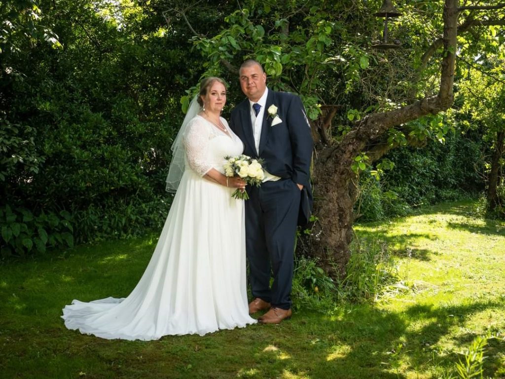 Photo of one of our real Lucky Sixpence brides and her new husband standing in a grassy area by a tree.