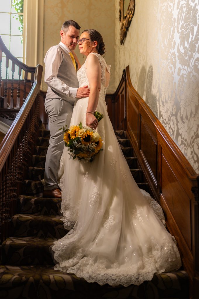Photo of one of our real Lucky Sixpence brides. She is standing on a set of stairs with her new husband holding a bouquet of yellow sunflowers.
