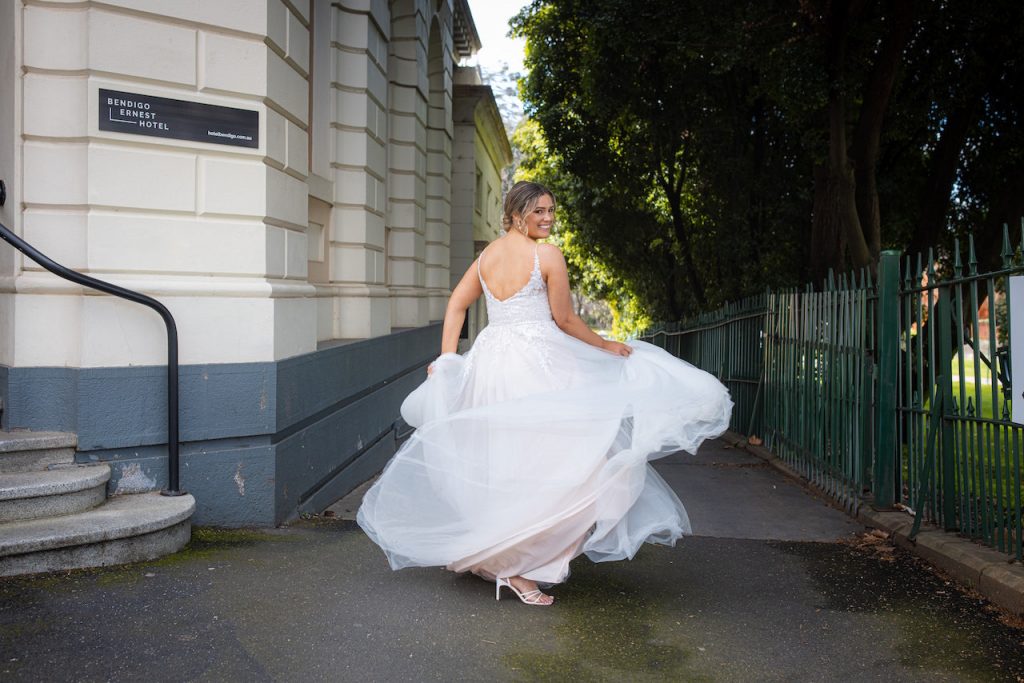 Photo shows a bride spinning in a wedding dress. She has her back to the camera and she is looking over her right shoulder towards the camera. The dress is "Don't Stop" by Purple Fox Bride, and it features a lightly sequinned bodice and plain tulle skirt.