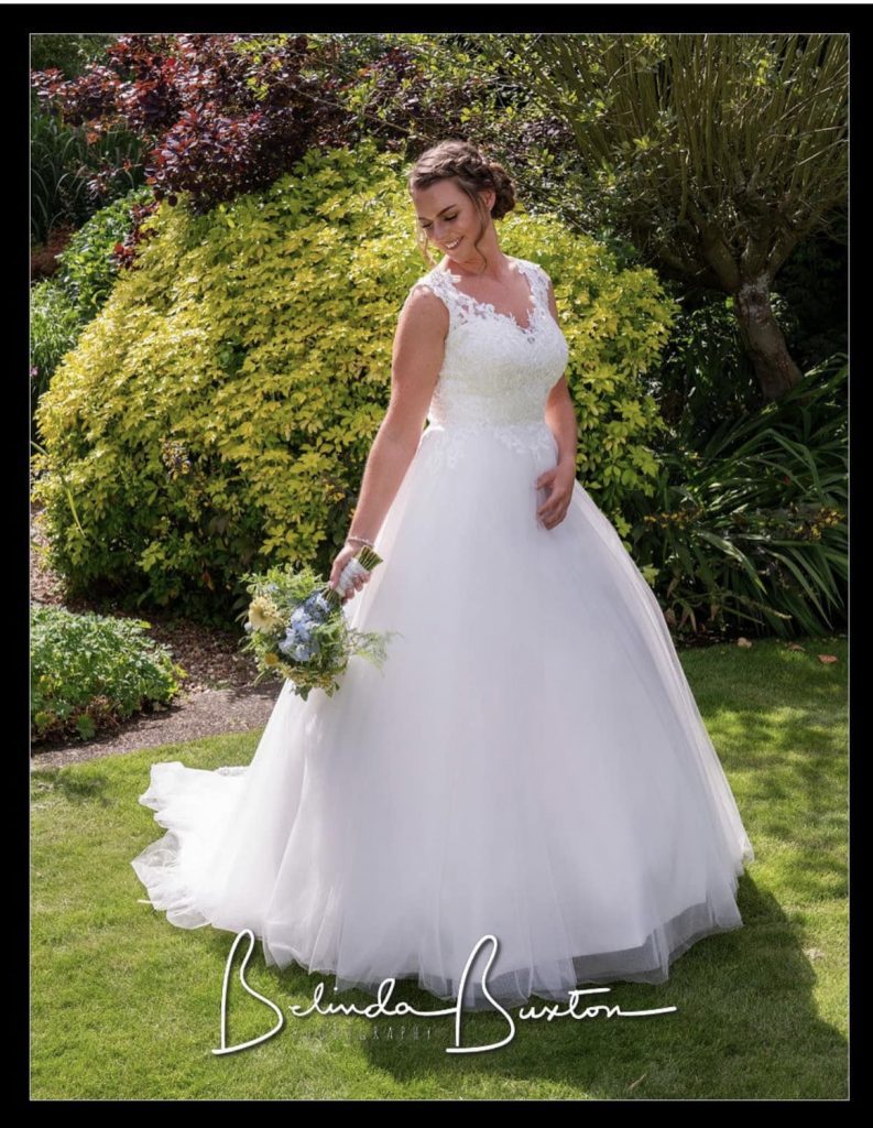 Photo shows real bride Emily standing on grass in some gardens holding her bouquet. She is wearing a Hilary Morgan ivory wedding dress.