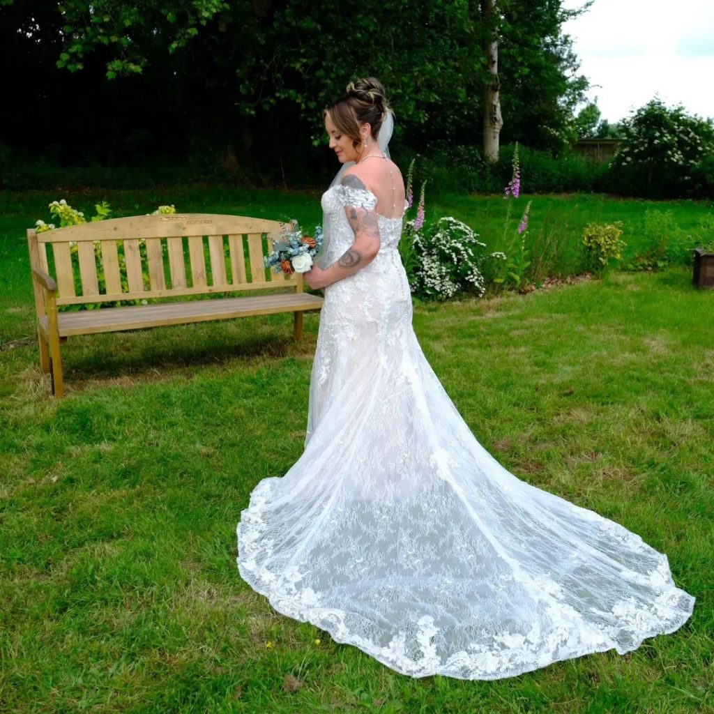 Real bride Kirsty standing on grass in front of a bench showing the gorgeous train on her lace fitted dress.