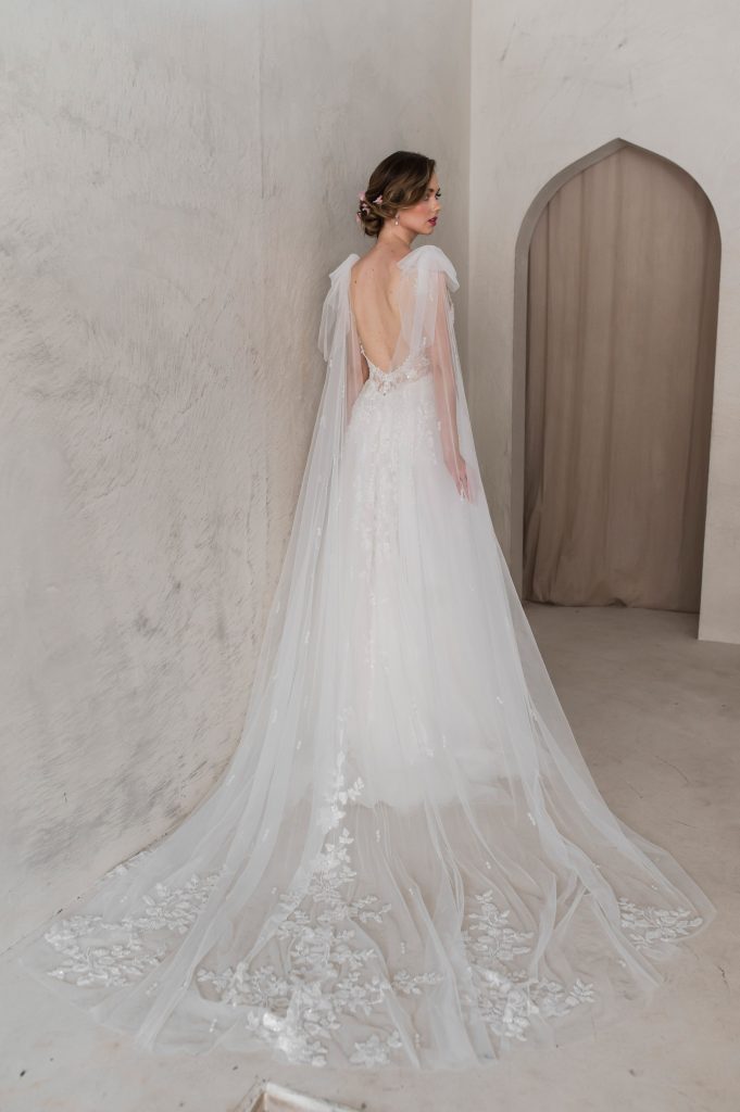 The Lucky Sixpence Bridal guide to veils. Photo shows a bride with her back to the camera. She is looking over her right shoulder. She is wearing a lace and tulle A-line bridal gown with a tulle cape veil cascading from her shoulders. There is beading at the end of the cape.