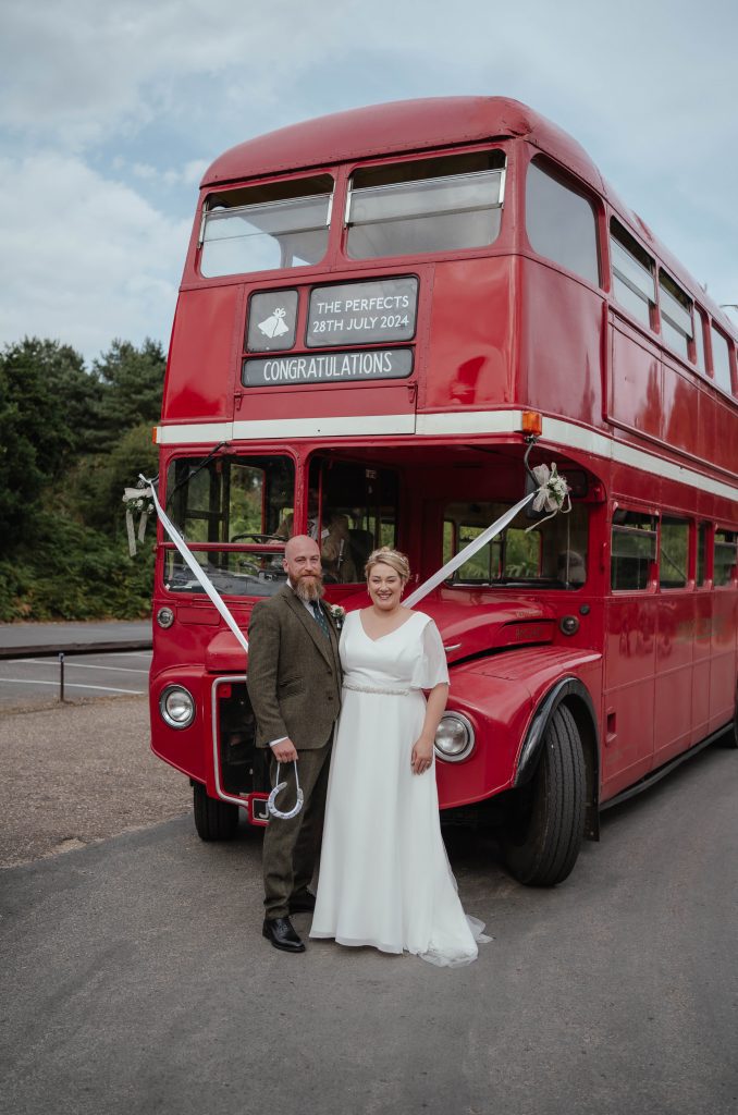Lucky Sixpence Bridal real bride. Photo shows real bride Charlotte. Bride and groom are standing in front of a red London double-decker bus.