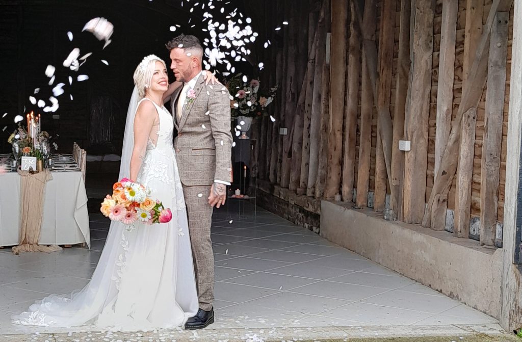 The Lucky Sixpence Bridal guide to veils. Photo shows bride and groom standing in the front of an old barn. Bride has her arm around the groom and is holding her bouquet in her other hand. Confetti is showering down.