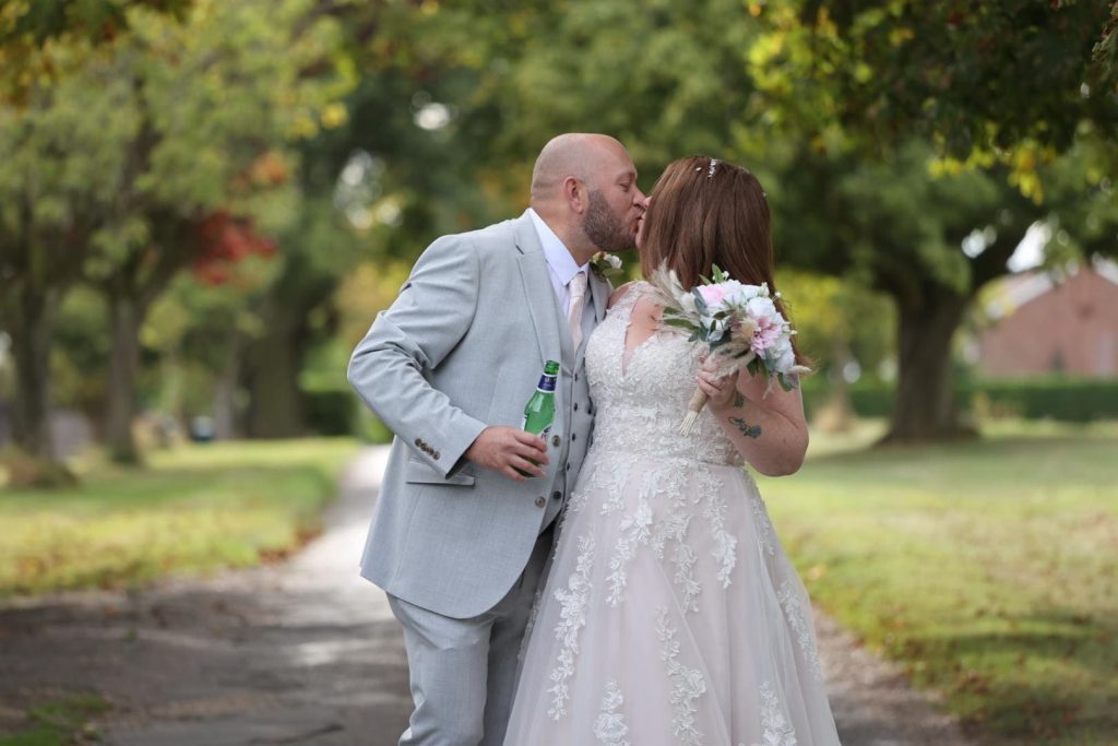 Lucky Sixpence Bridal real bride. Photo shows real bride Amy. Bride and groom are kissing. Bride is holding her bouquet and groom is holding a bottle of beer. There are trees in the background. Photo taken by Pitcher Perfect Events.