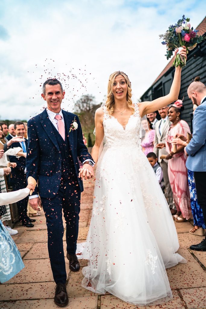 Lucky Sixpence Bridal real bride, A. Photo shows smiling bride and groom covered in confetti. Bride is holding her bouquet aloft while their guests look on.