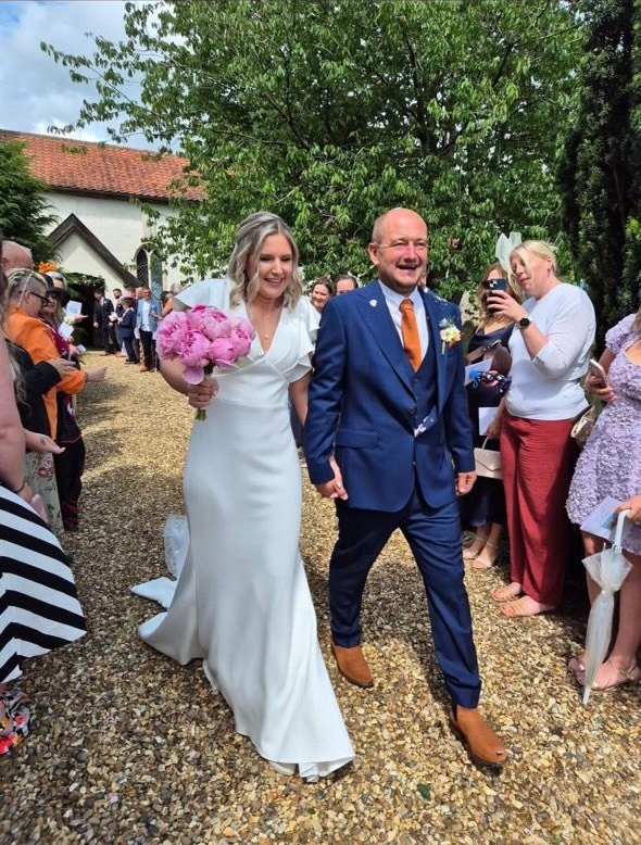 Photo shows a Lucky Sixpence bride holding her new husband's hand, walking past a line of guests wearing a fitted gown with flutter sleeves and holding a bouquet of pink peonies.