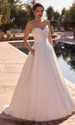 Photo shows bride standing poolside looking at the camera, with her hands in the pockets of a strapless sweetheart neckline bridal gown.