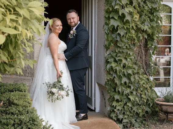 Photo shows a real Lucky Sixpence bride standing in a doorway. Her new husband has his hands on her waist, and they are both looking at the camera. The bride is also holding a bouquet of spring flowers.