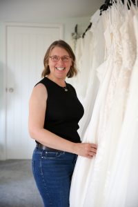 Photograph shows Vicky, the owner of Lucky Sixpence Bridal, standing in front of a rail of wedding dresses. She has chin length bobbed hair and brown rimmed glasses, and she is wearing jeans and a black sleeveless top.