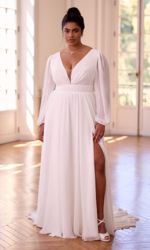 Photo shows model standing in an open room with windows behind her. She is wearing Juliana by Bianco Evento, a long chiffon wedding dress in ivory, with long sleeves gathered at the wrist, a plunge neckline, wide waistband, and gathered skirt with leg split.