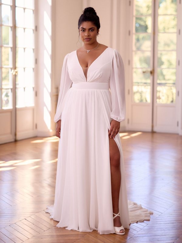 Photo shows model standing in an open room with windows behind her. She is wearing Juliana by Bianco Evento, a long chiffon wedding dress in ivory, with long sleeves gathered at the wrist, a plunge neckline, wide waistband, and gathered skirt with leg split.