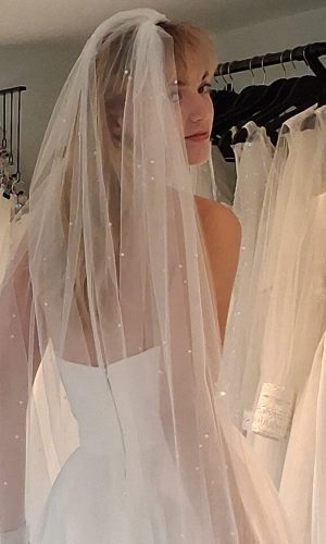 Photo shows model looking over her right shoulder at the camera wearing an ivory cut-edge veil with scatter pearls.