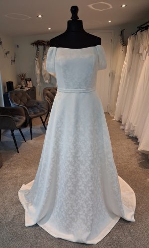 Photo shows and ivory jacquard wedding dress on a mannequin. The dress has a slight scoop neckline, off-the-shoulder straps, and belt.