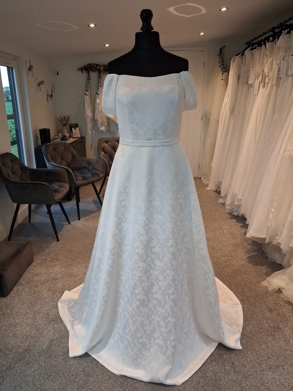 Photo shows and ivory jacquard wedding dress on a mannequin. The dress has a slight scoop neckline, off-the-shoulder straps, and belt.